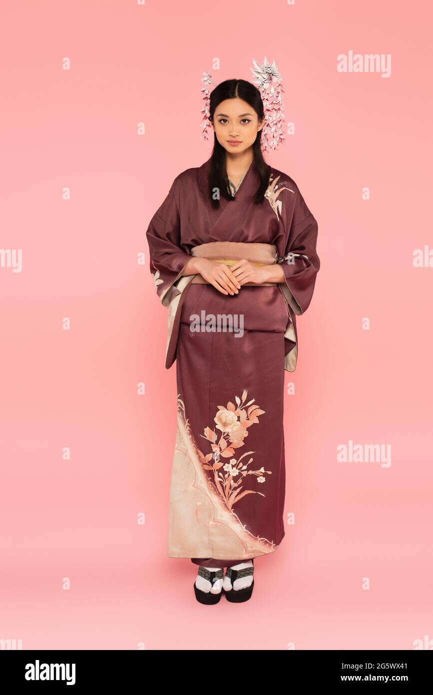 In voller Länge eine asiatische Frau in Kimono und Flip-Flops, die auf rosa Hintergrund die Kamera anschaut Stockfoto