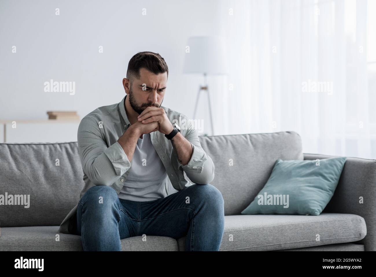 Nachdenklicher junger, seriös-gutaussehender Mann sitzt zu Hause auf dem Sofa, in Gedanken verloren und denkt über Problemlösung nach Stockfoto