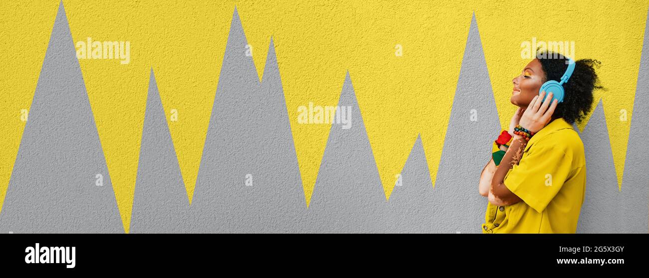 Musikliebhaberin hört Musik von ihren blauen Kopfhörern in der Nähe einer Stadtwand, die in den Farben 2021 Ultimate Grey und Illuminating Yellow mit gemalt ist Stockfoto