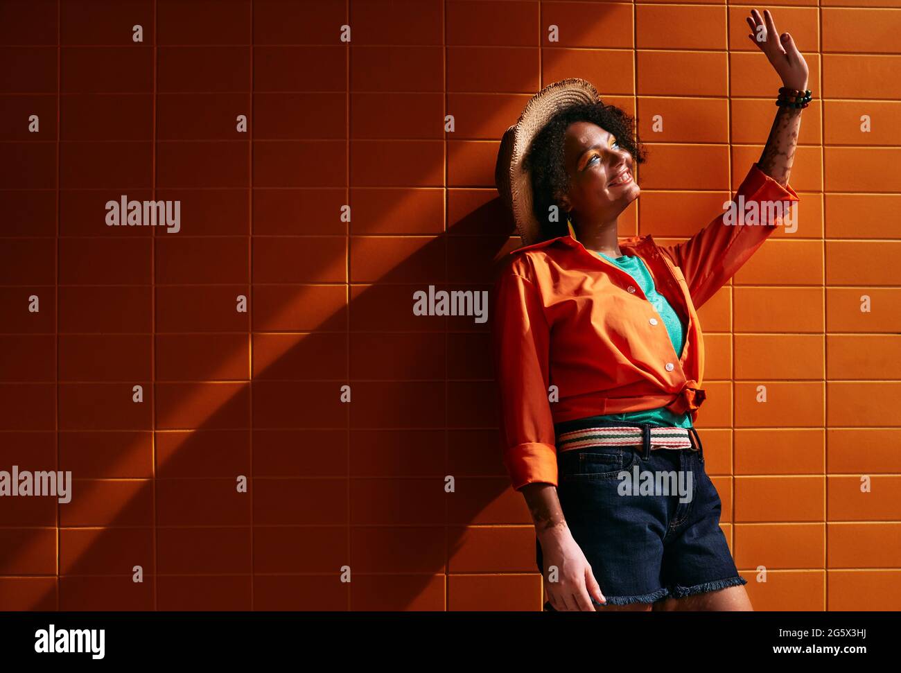 Lächelnde afroamerikanische Frau in trendigen Kleidern und Strohhut an der orangefarbenen Gebäudewand steht in Sonnenstrahlen mit langem Schatten Stockfoto