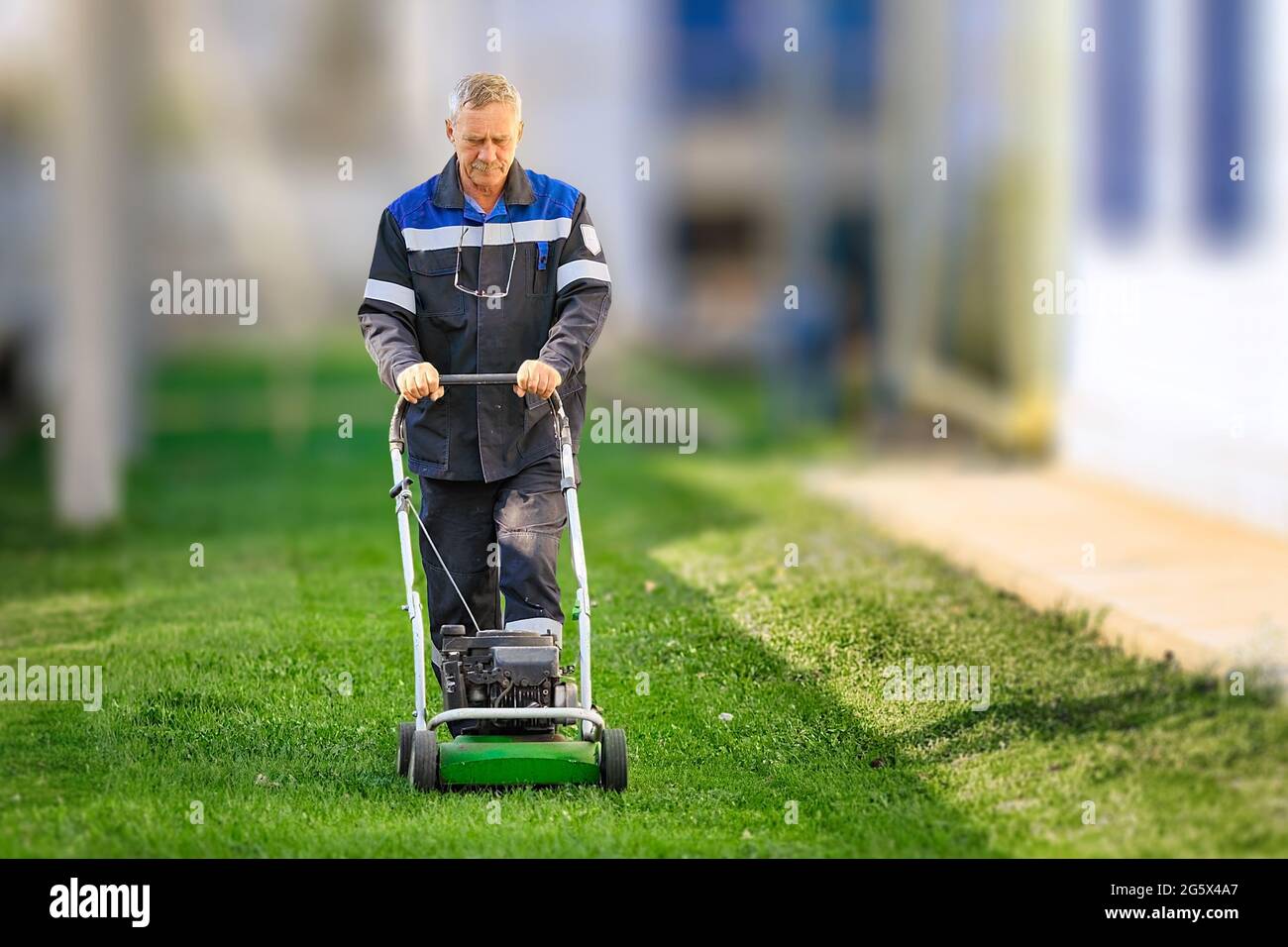 Ein älterer Arbeiter mäht an einem sonnigen Tag den Rasen. Ein Mann mit Arbeitskleidung arbeitet mit einem Rasenmäher auf einer grünen Wiese. Vorderansicht. Stockfoto