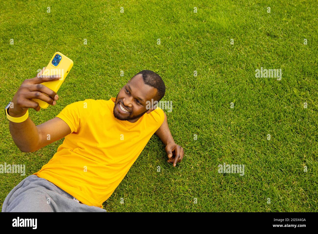Fröhlicher, hübscher afro-amerikanischer Mann, der auf dem Gras liegt, sich entspannt und nach dem Sport eine Pause macht. Lächelnder schwarzer Kerl, der online mit spricht Stockfoto