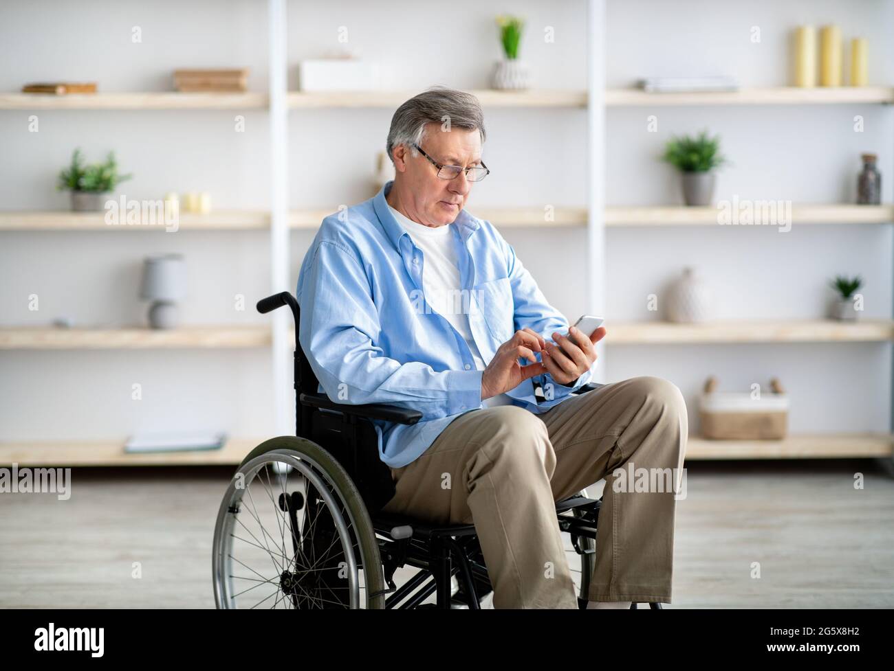 Älterer behinderter Mann im Rollstuhl, der Smartphone benutzt, Nachrichten überprüft, Videos ansieht, zu Hause im Internet surft Stockfoto