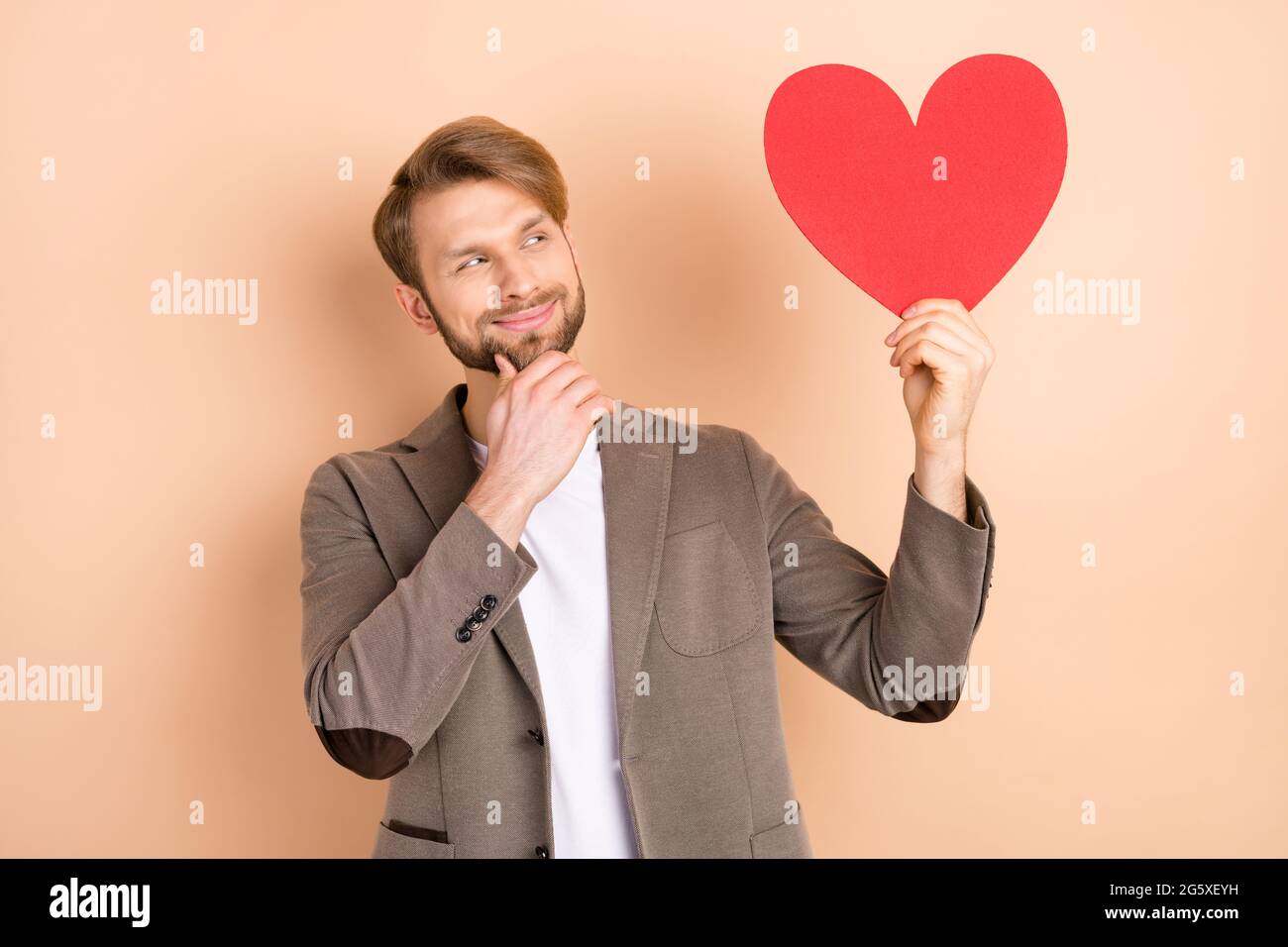 Foto von nachdenklich ziemlich Gentleman gekleidet Blazer Arm Kinn halten suchen große rote Herz leeren Raum isoliert beige Farbe Hintergrund Stockfoto