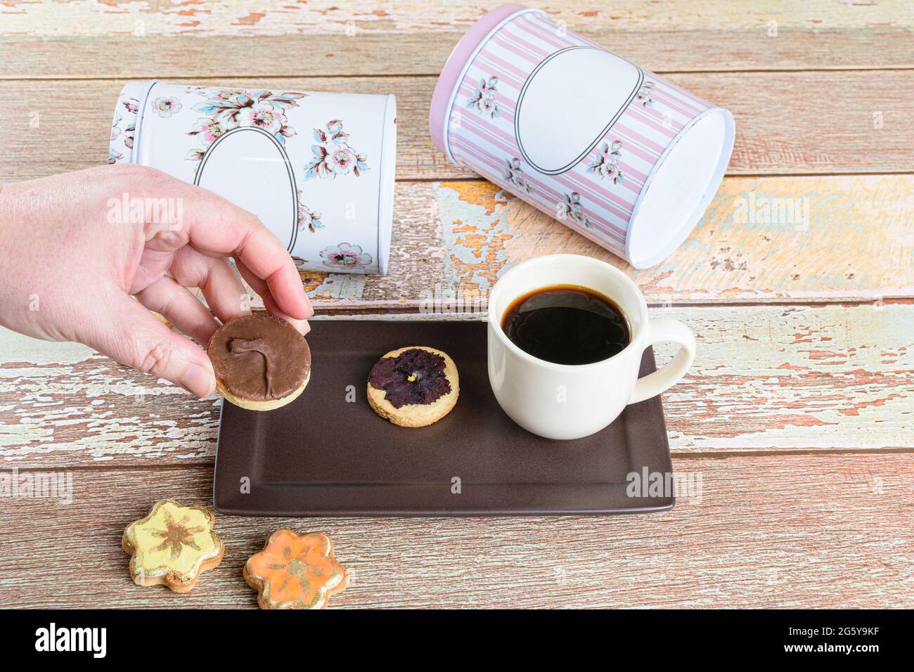 Frau fängt einen mit Schokolade überzogenen butterigen Keks, neben anderen Keksen, einer Tasse Kaffee und Dosen, die sich hinlegen. Stockfoto