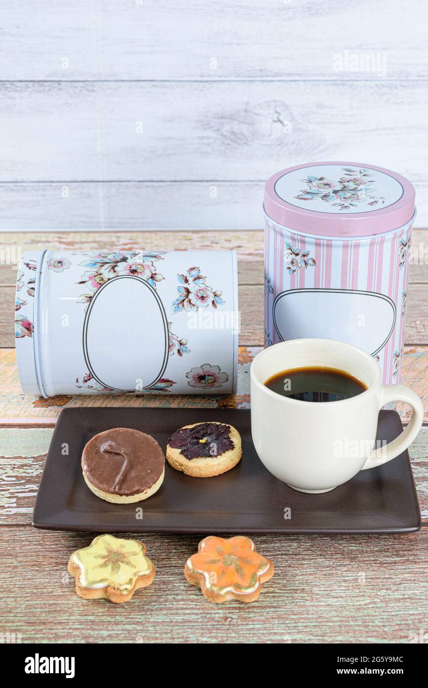 Buttergebäck mit einer dehydrierten Blume oben, neben anderen Keksen, einer Tasse Kaffee und Dosen (vertikales Foto). Stockfoto