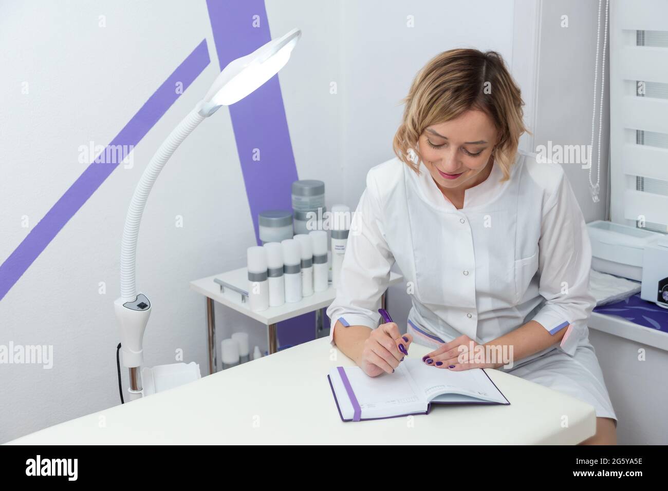 Junge Kosmetologin, die Untersuchungsergebnisse aufzeichnet und in einem modernen Kosmetikbüro lächelt Stockfoto