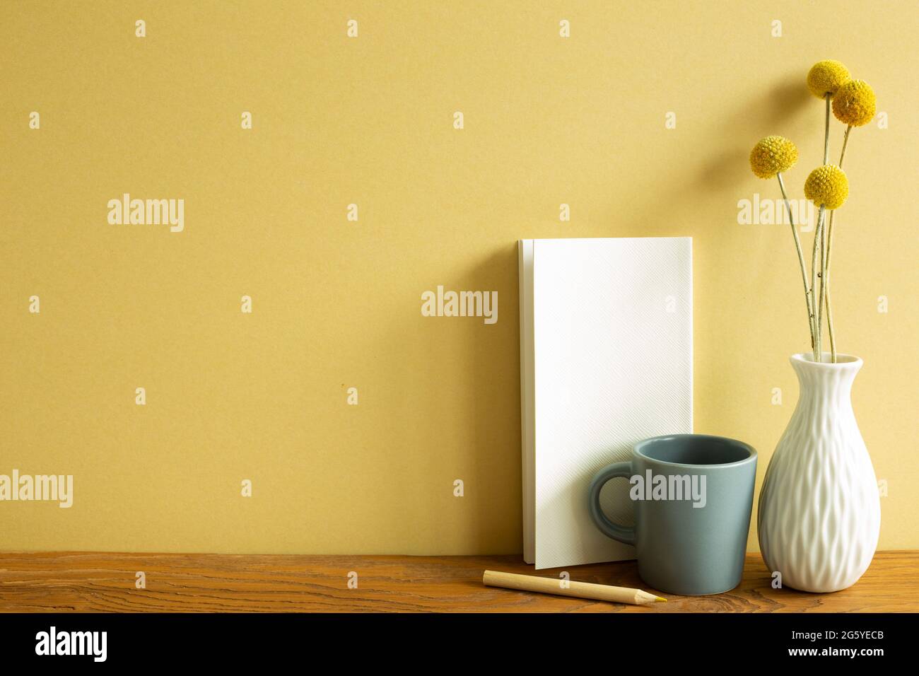 Notizbuch, Becher, Bleistift, Vase mit trockenen Blumen auf Holzschreibtisch. Gelber Wandhintergrund. Arbeitsbereich Stockfoto