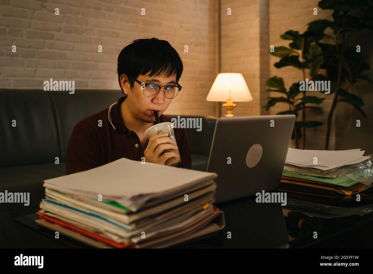 Gestresster asiatischer Geschäftsmann in legerer Kleidung, der bis spät in die Nacht mit Akte und Laptop arbeitet, während er Kaffee trinkt, um wach zu bleiben und sich zu Hause zu konzentrieren Stockfoto