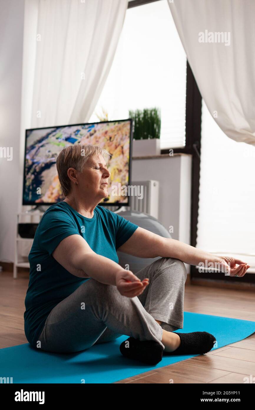 Friedliche ältere Frau, die bequem in Lotusposition auf einer Yogamatte sitzt und mit geschlossenen Augen Meditation praktiziert. Ruhiger Rentner, der Fitness trainiert, Körperübungen während des Meditationstrainings im Wohnzimmer Stockfoto