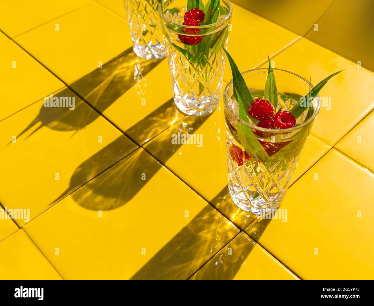 Erfrischende Sommergetränke in Kristallgläsern mit grünen Blättern Himbeer-gelbe Kacheln Hintergrund Schatten. Süßer, kalter natürlicher Cocktail an heißen Sommertagen. So Stockfoto
