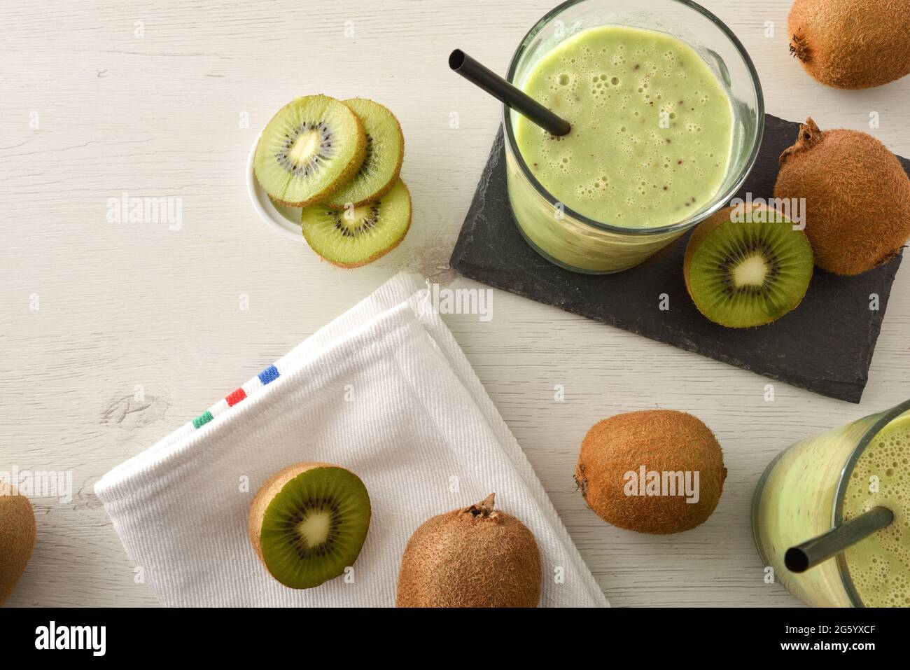 Gläser mit Kiwi-Drink auf Schieferteller auf weißer Holzbank voller Früchte mit weißer Serviette. Draufsicht. Stockfoto