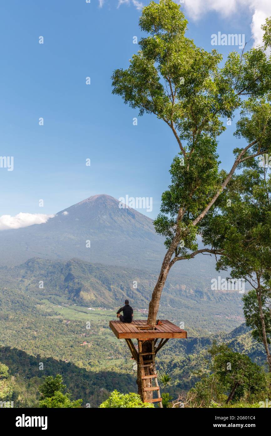 Mann, der auf einer Baumplattform an einem Aussichtspunkt mit Blick auf den Mount Agung (Gunung Agung) sitzt, Vulkan in Karangasem Regency, Bali, Indonesien Stockfoto