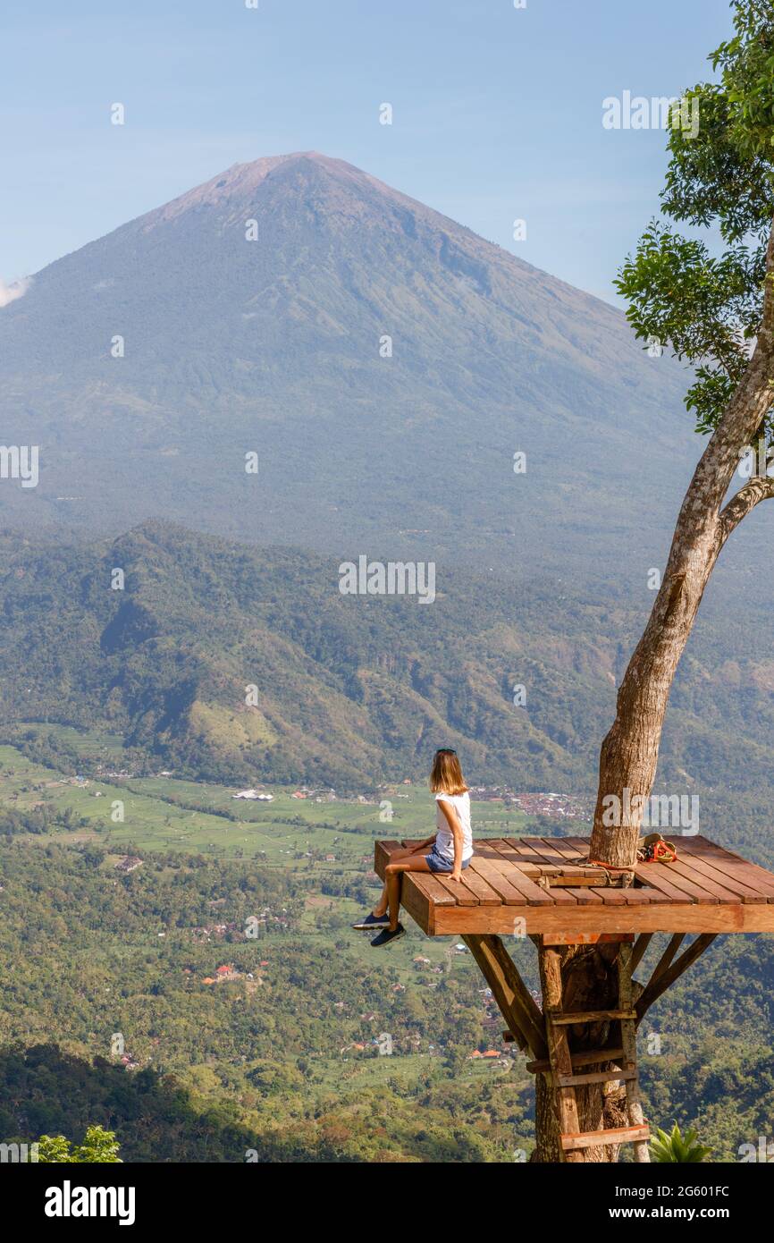 Ein Mädchen, das auf einer Baumplattform an einem Aussichtspunkt mit Blick auf den Mount Agung (Gunung Agung), Vulkan in Karangasem Regency, Bali, Indonesien, sitzt Stockfoto