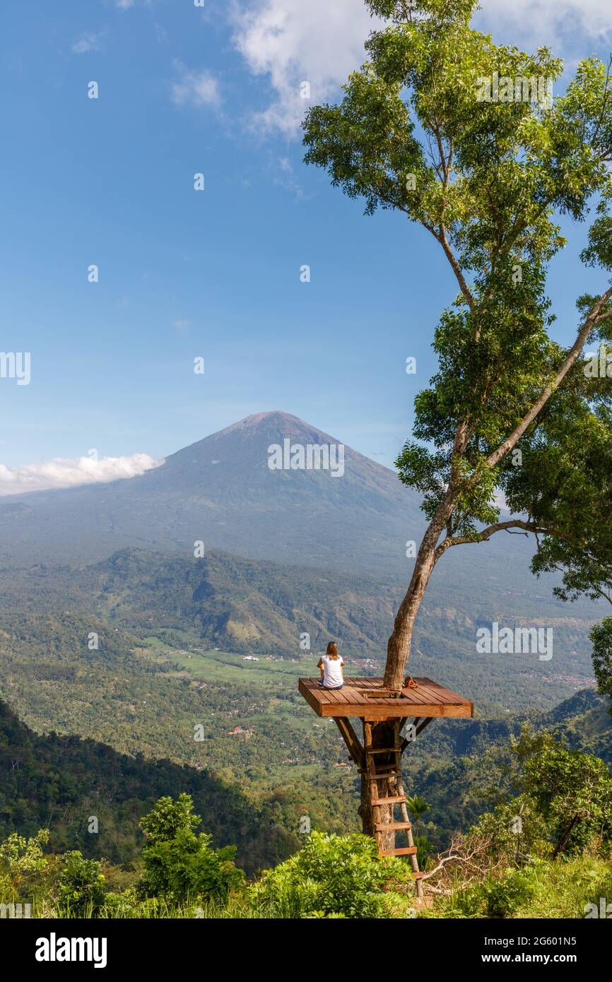 Ein Mädchen, das auf einer Baumplattform an einem Aussichtspunkt mit Blick auf den Mount Agung (Gunung Agung), Vulkan in Karangasem Regency, Bali, Indonesien, sitzt Stockfoto