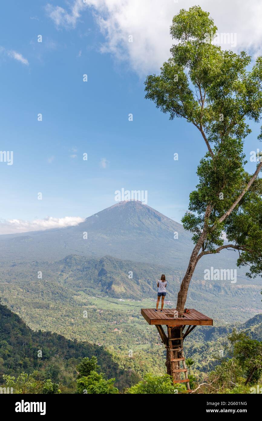 Ein Mädchen, das auf einer Baumplattform an einem Aussichtspunkt mit Blick auf den Mount Agung (Gunung Agung), Vulkan in Karangasem Regency, Bali, Indonesien, steht Stockfoto