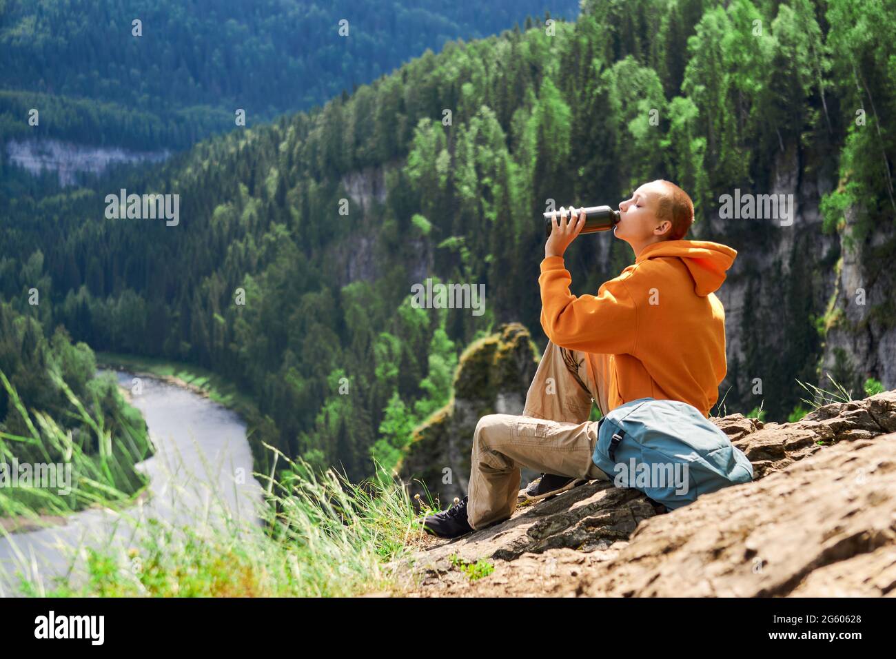 Teenager-Wandermädchen ruhen sich aus und trinken am Rand einer Klippe mit Blick auf den Fluss vor dem Hintergrund einer natürlichen Landschaft Stockfoto