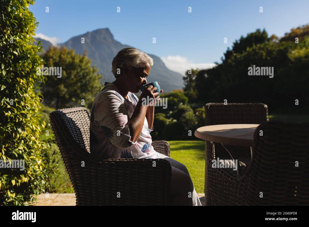 Ältere afroamerikanische Frau, die in einem sonnigen Garten Tee trinkt, atemberaubende Landschaft Stockfoto
