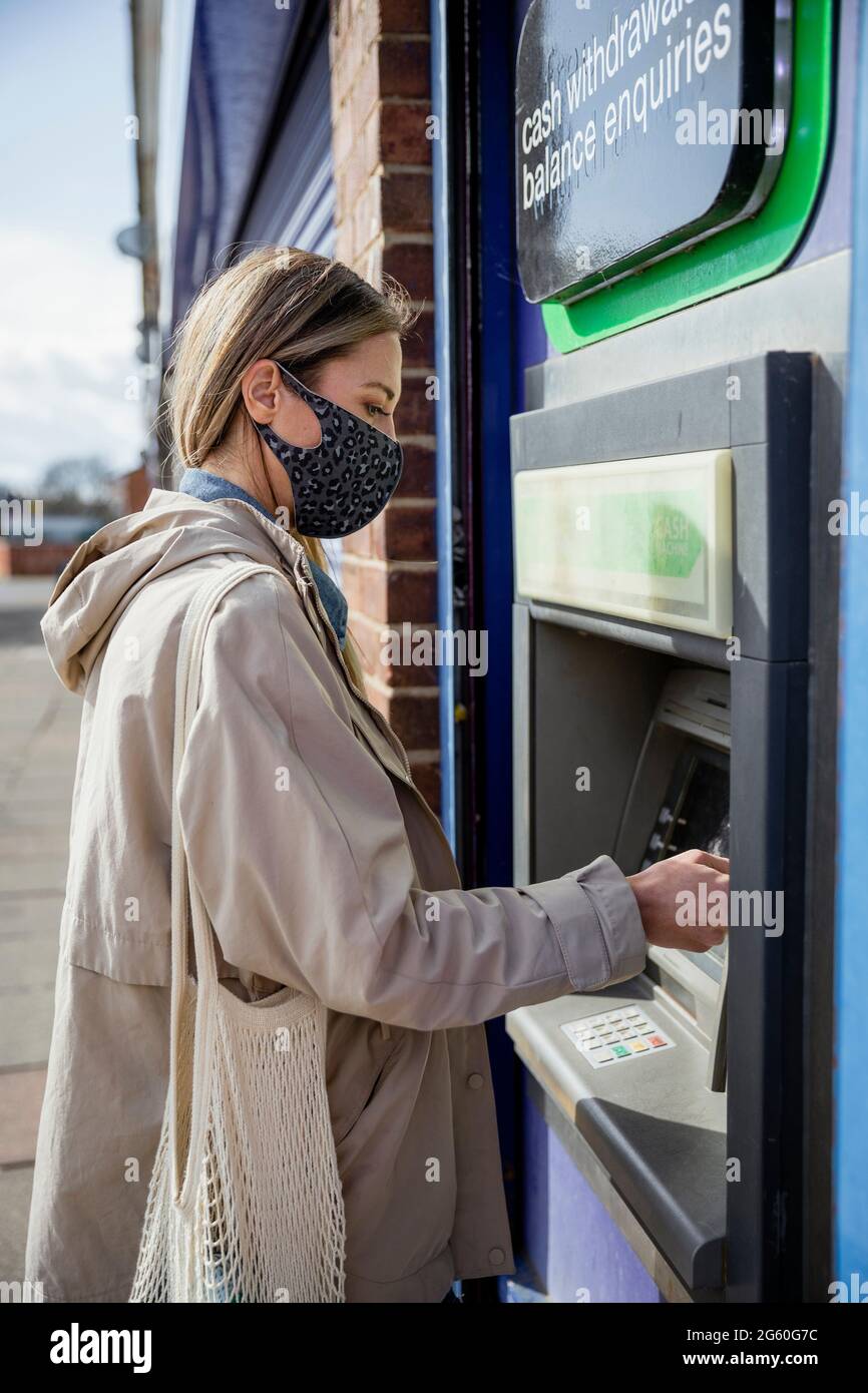 Eine Seitenansicht einer kaukasischen Frau mit mittlerem Erwachsenen, die an einem Geldautomaten auf einer Stadtstraße eine Debitkarte verwendet, trägt lässige Kleidung und ein schützendes fa Stockfoto