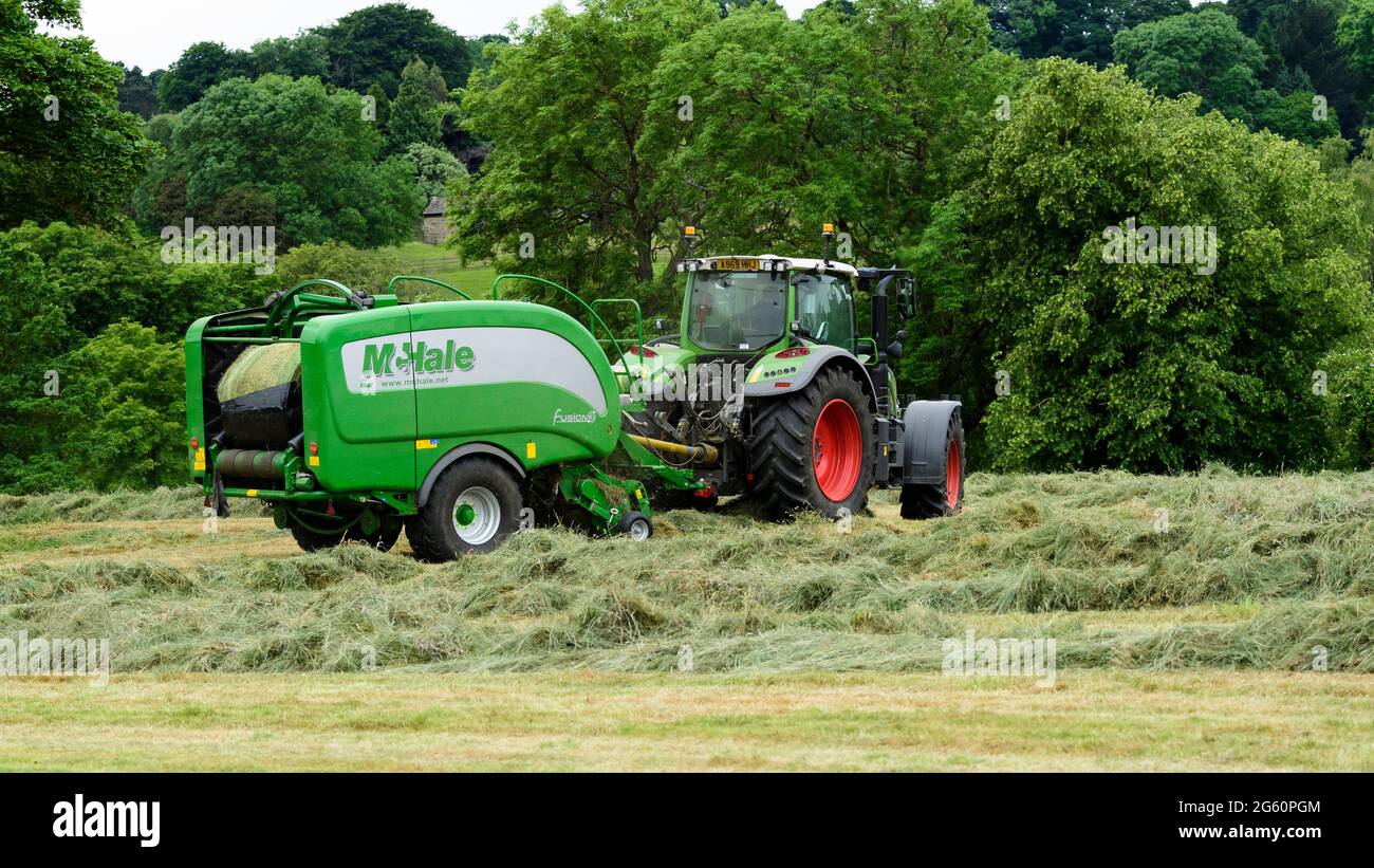 Heu- oder Silageherstellung (Landwirt im Traktor auf dem Bauernhof bei der Arbeit auf dem Land, beim Sammeln von trockenem Gras, beim Einwickeln von Rundballen in Ballenpressen) - Yorkshire England, Großbritannien. Stockfoto