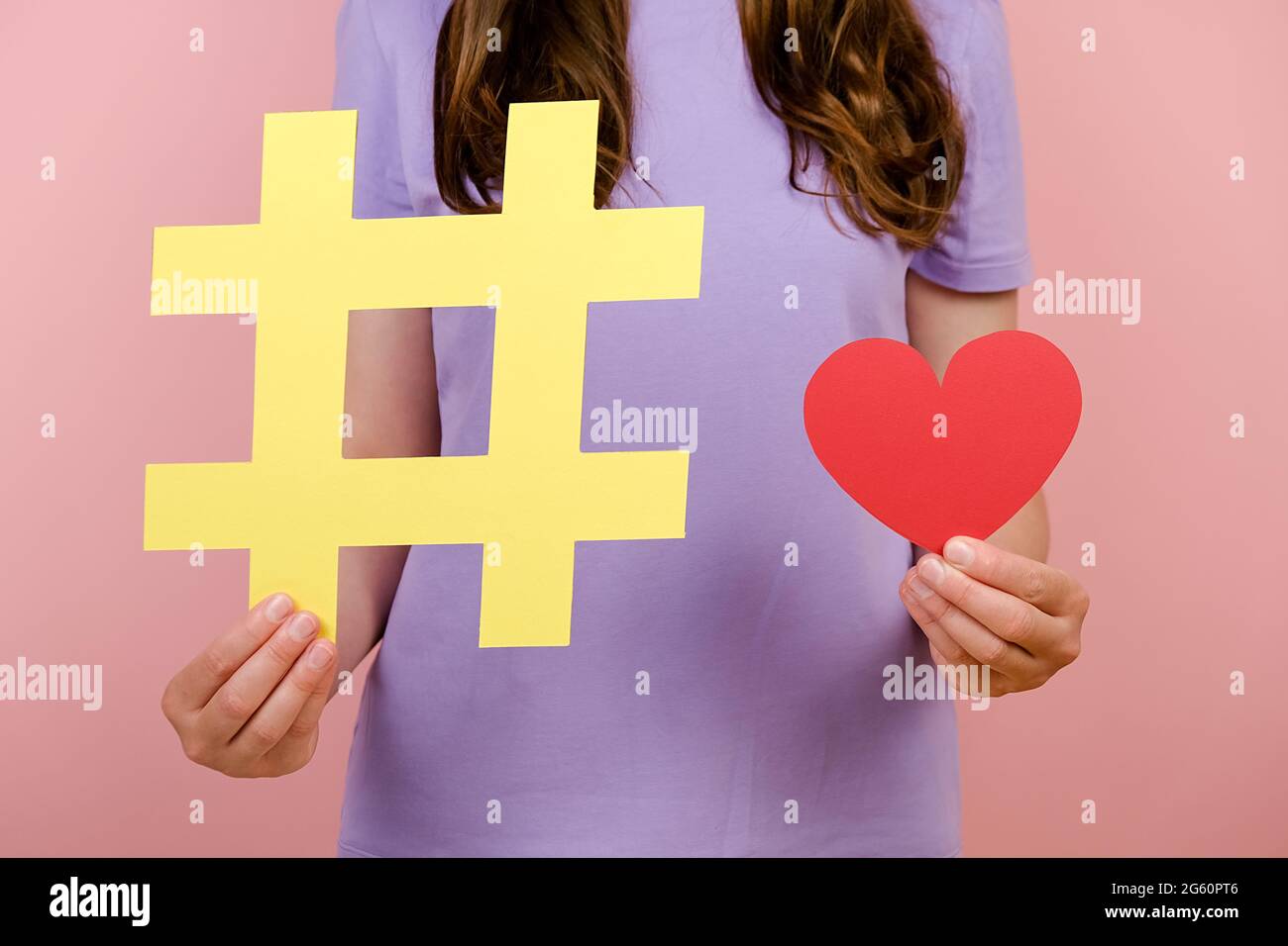Nahaufnahmen von zugeschnittenen Aufnahmen einer jungen Frau, die ein T-Shirt trägt, zeigen ein großes gelbes Hashtag-Schild und ein kleines rotes Herz, das isoliert über einer rosa Wand posiert Stockfoto