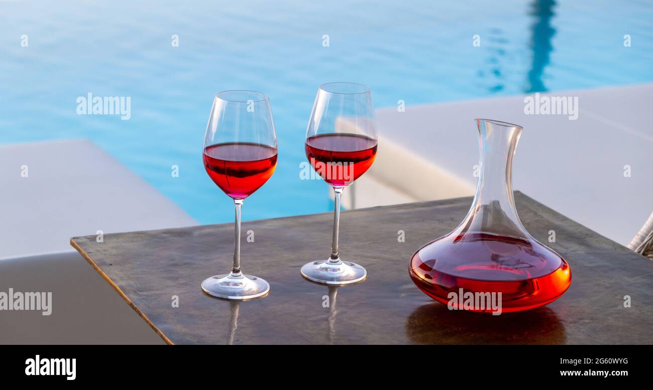 Rotweingläser am Swimmingpool. Zwei Weingläser und Dekanter auf Holztisch, türkisblauer Wasserhintergrund. Entspannung und Romantik durch die RE Stockfoto
