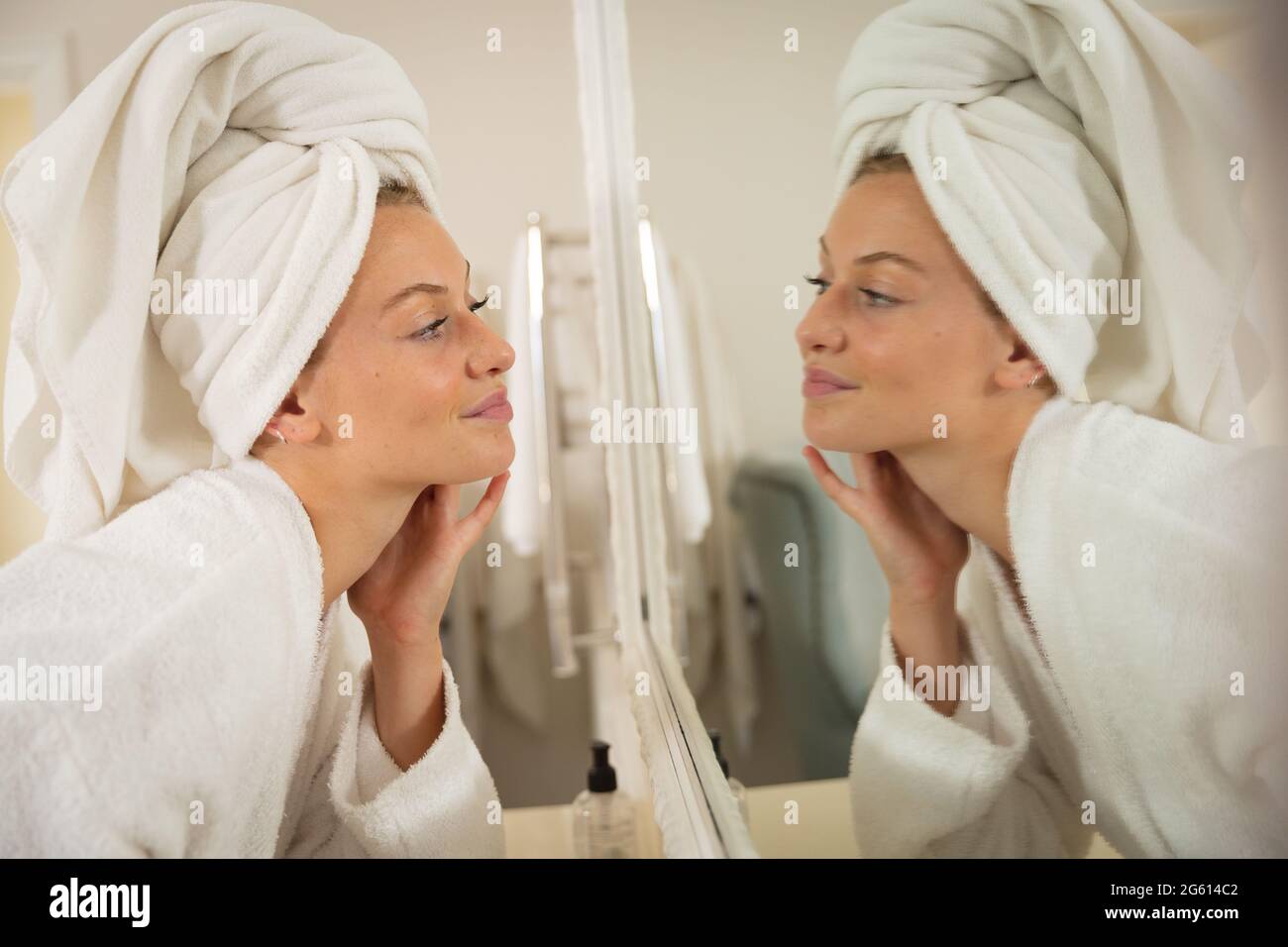 Lächelnde kaukasische Frau im Badezimmer mit Handtuch auf dem Kopf, Blick in Spiegel und feuchtigkeitsspendende Gesicht Stockfoto