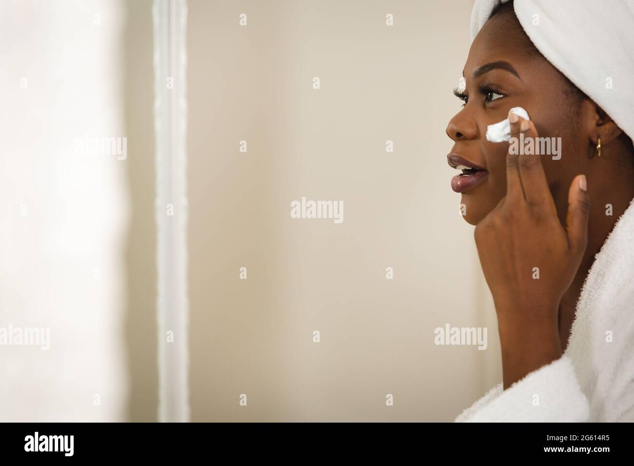 afroamerikanische Frau im Badezimmer mit Handtuch auf dem Kopf, Blick in Spiegel feuchtigkeitsspendende Gesicht Stockfoto