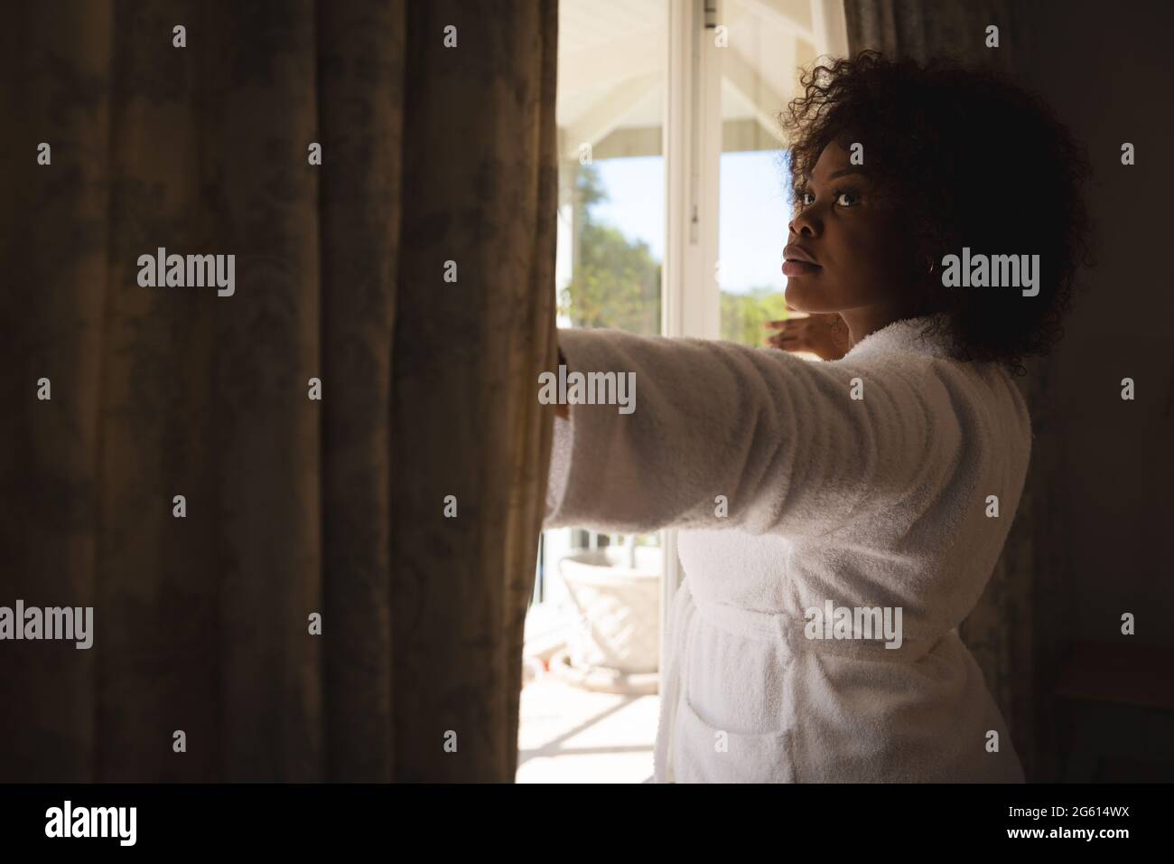 afroamerikanische Frau, die an einem sonnigen Fenster steht und Vorhänge zieht und wegschaut Stockfoto