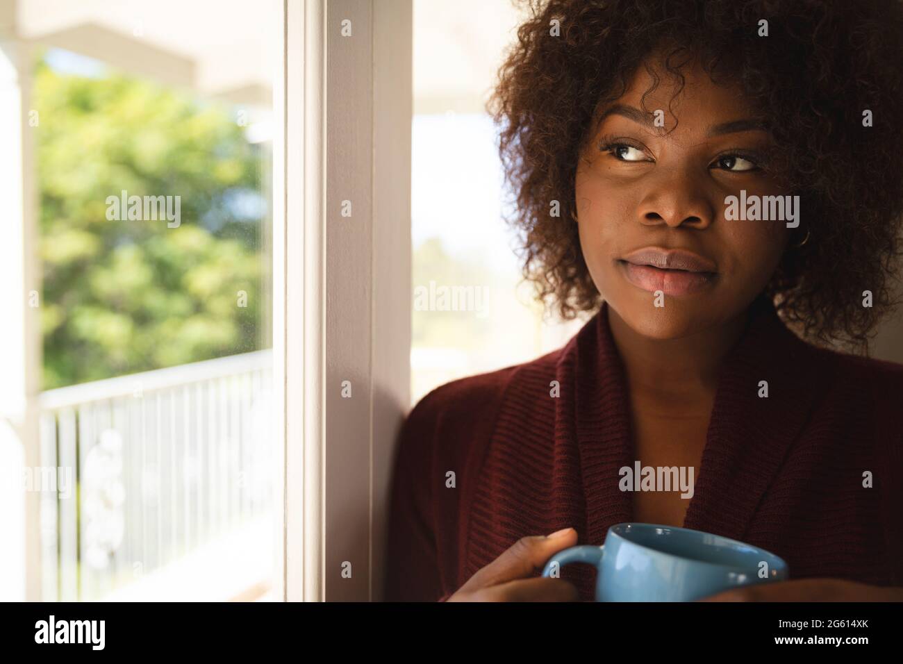 Nachdenkliche afroamerikanische Frau, die aus dem sonnigen Fenster blickt und eine Tasse Kaffee hält Stockfoto