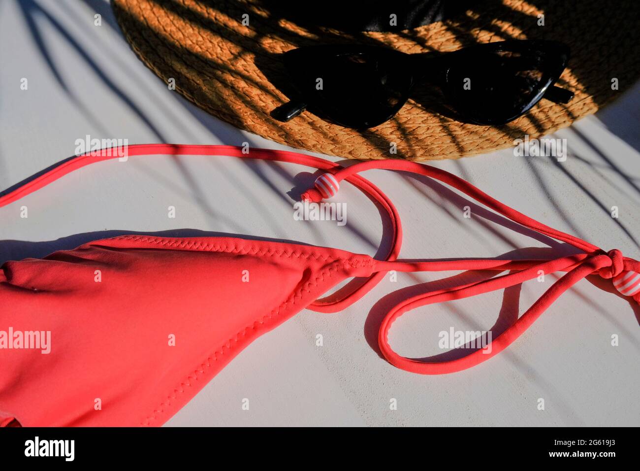 Sonnenbrille auf dem Strohhut, rotes Bikinitop in der Handfläche hinterlässt Schatten auf weißem Hintergrund. Sommer flach liegend. Urlaubskonzept Stockfoto