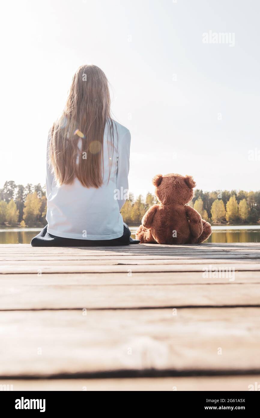 Kleines Mädchen mit Teddybär sitzt am Pier in der Nähe des ruhigen Flusses Stockfoto
