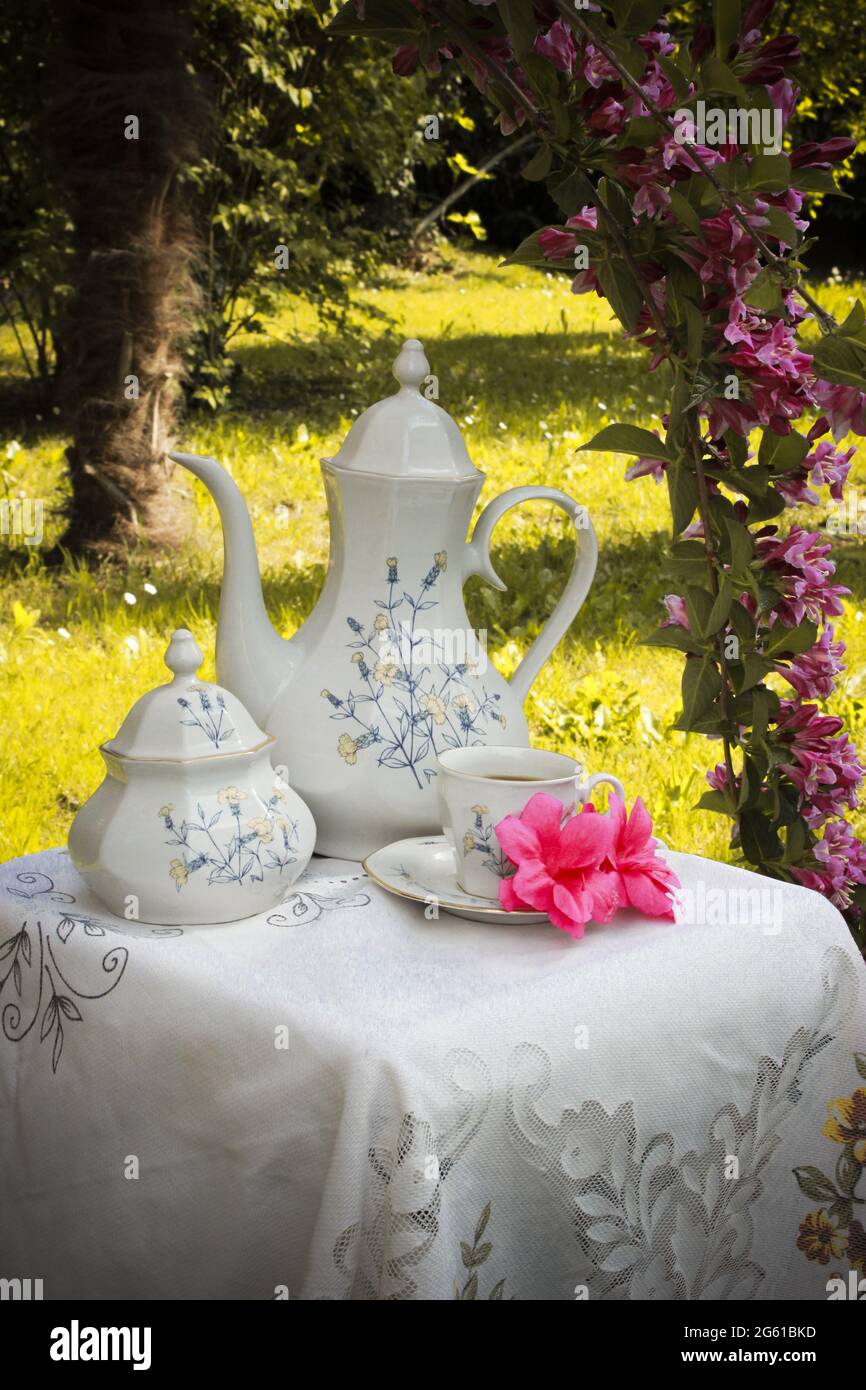 Tisch mit antiker Teekannen, Tasse und Zuckerdose mit einem herrlichen Garten voller Blumen im Hintergrund im Licht des Sonnenuntergangs Stockfoto