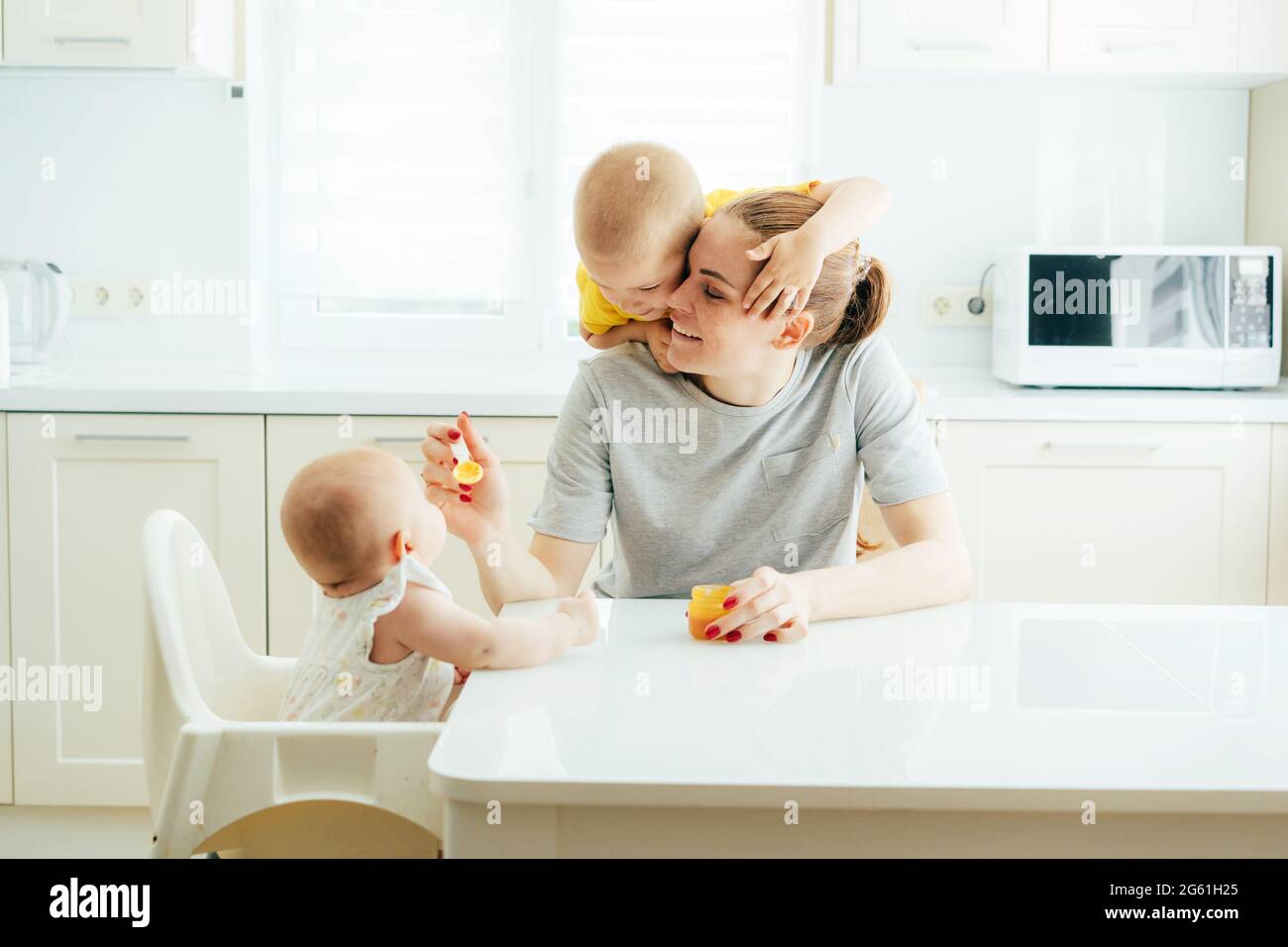 Familienspaß, Mama füttert die Kinder in der Küche. Mama füttert das Baby mit falschem Gemüsepüree, das ältere Kind umarmt die Mutter. Momente des Familienvergnügens Stockfoto