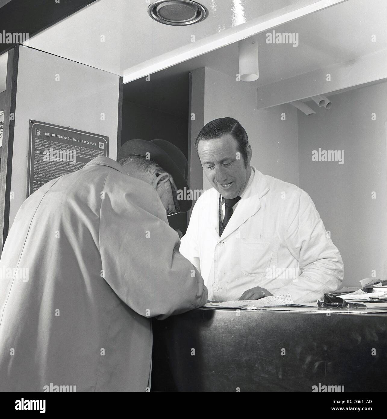 1975, historisch, ein weiß beschichteter Berater für Garagendienste, der hinter einem Verkaufstheke steht, wo ein älterer männlicher Kunde seine Unterschrift auf einige Papiere legt, England, Großbritannien. Stockfoto