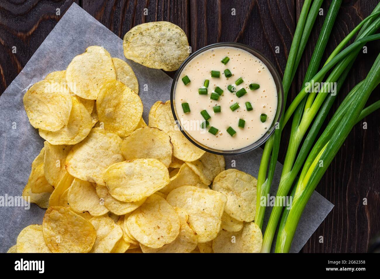Knusprige Kartoffelchips, ein paar frische grüne Zwiebeln und Mayonnaise auf einem schwarzen Holztisch. Snack aus frittierten dünnen Kartoffelscheiben. Ungesunde Ernährung. Stockfoto
