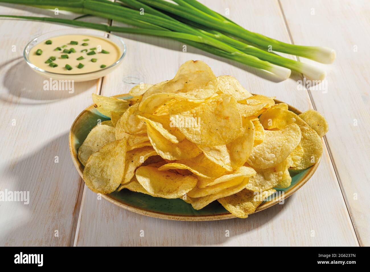 Knusprige Kartoffelchips auf einem Teller, ein paar frische grüne Zwiebeln und Mayonnaise auf einem Holztisch. Knuspriger Snack aus frittierten Kartoffelscheiben. Fast Food. Stockfoto