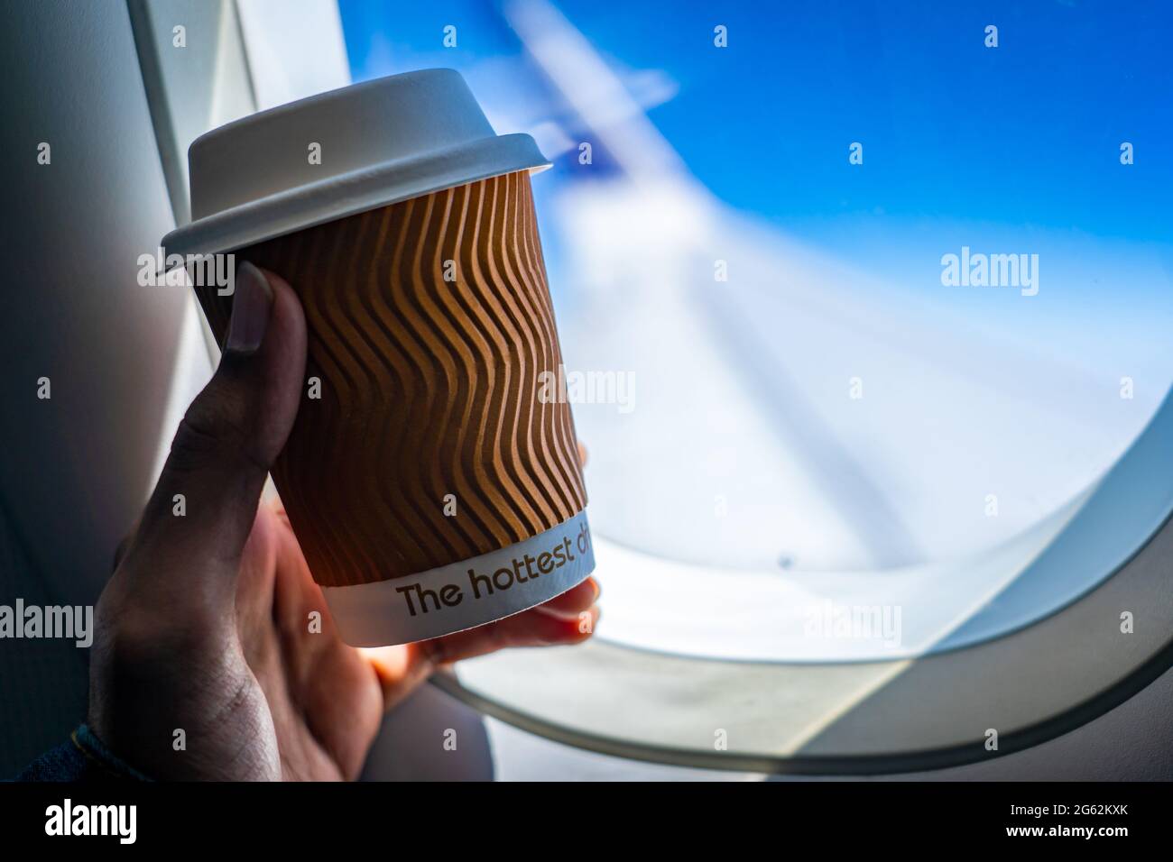 Selektiver Fokus auf eine recycelbare Kaffeetasse, die von einer männlichen linken Hand in der Nähe des Flugzeugfensters gehalten wird. Flugzeugflügel im Hintergrund verschwommen. Stockfoto