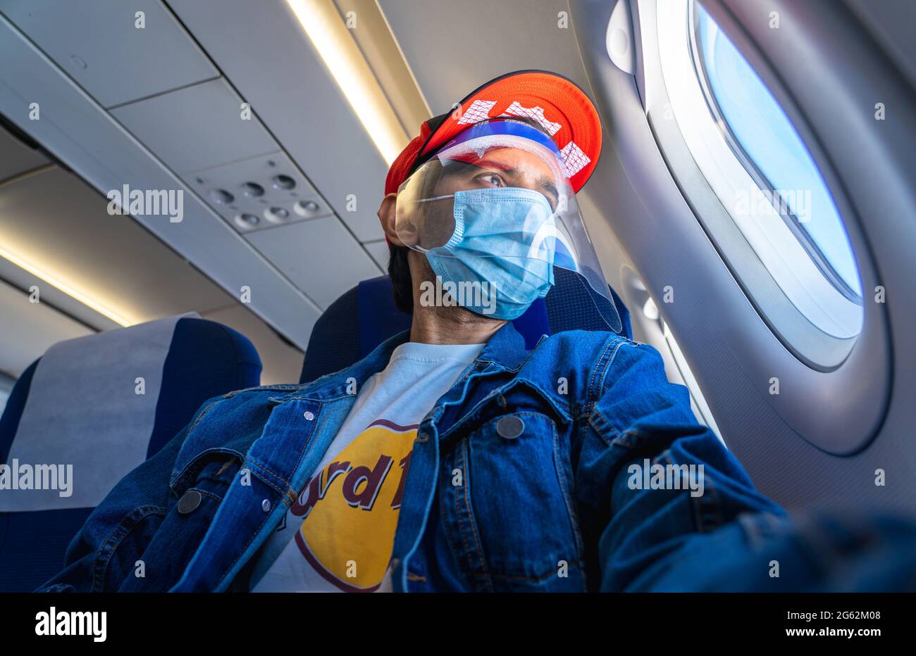 Ein junger Erwachsener, der eine Maske, einen Gesichtsschutz und einen roten Hut trägt, schaut vor das Flugzeugfenster. Stockfoto