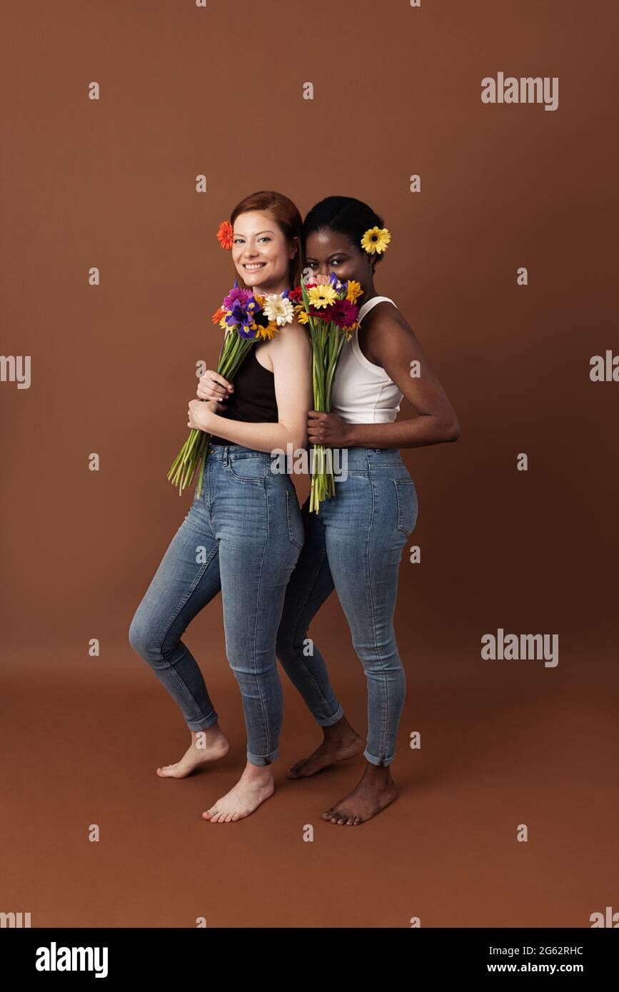 Zwei Frauen mit unterschiedlicher Hautfarbe posieren vor braunem Hintergrund. Fröhliche Weibchen mit Blumen, die zusammen stehen und die Kamera betrachten. Stockfoto