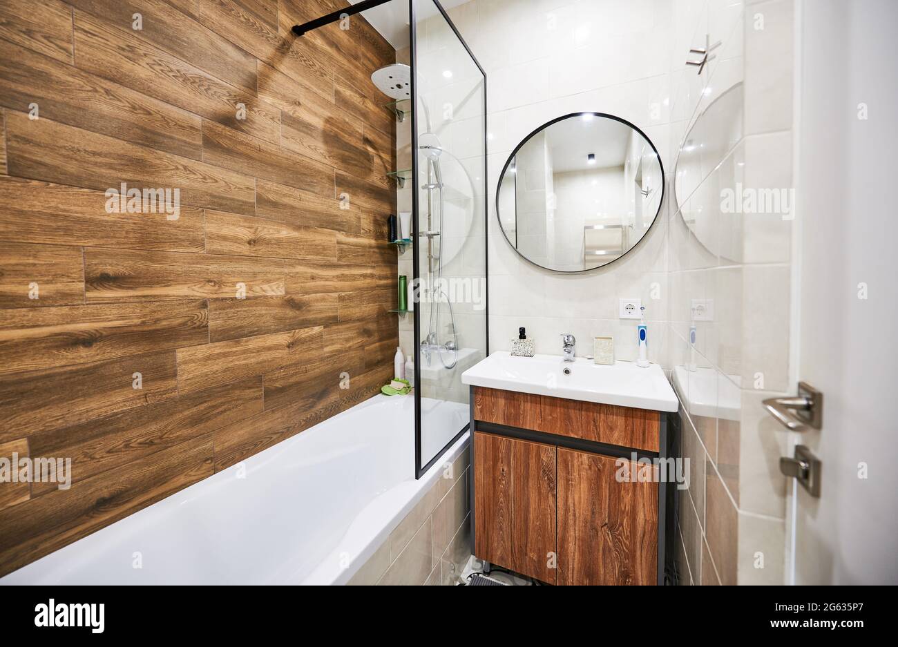 Stilvolle und komfortable Version des Baddesigns auf kleinem Raum. Weiße Farbe von rechteckiger Badewanne und Waschbecken in harmonischer Kombination mit rundem Spiegel, ergänzt durch Holzdekor. Stockfoto