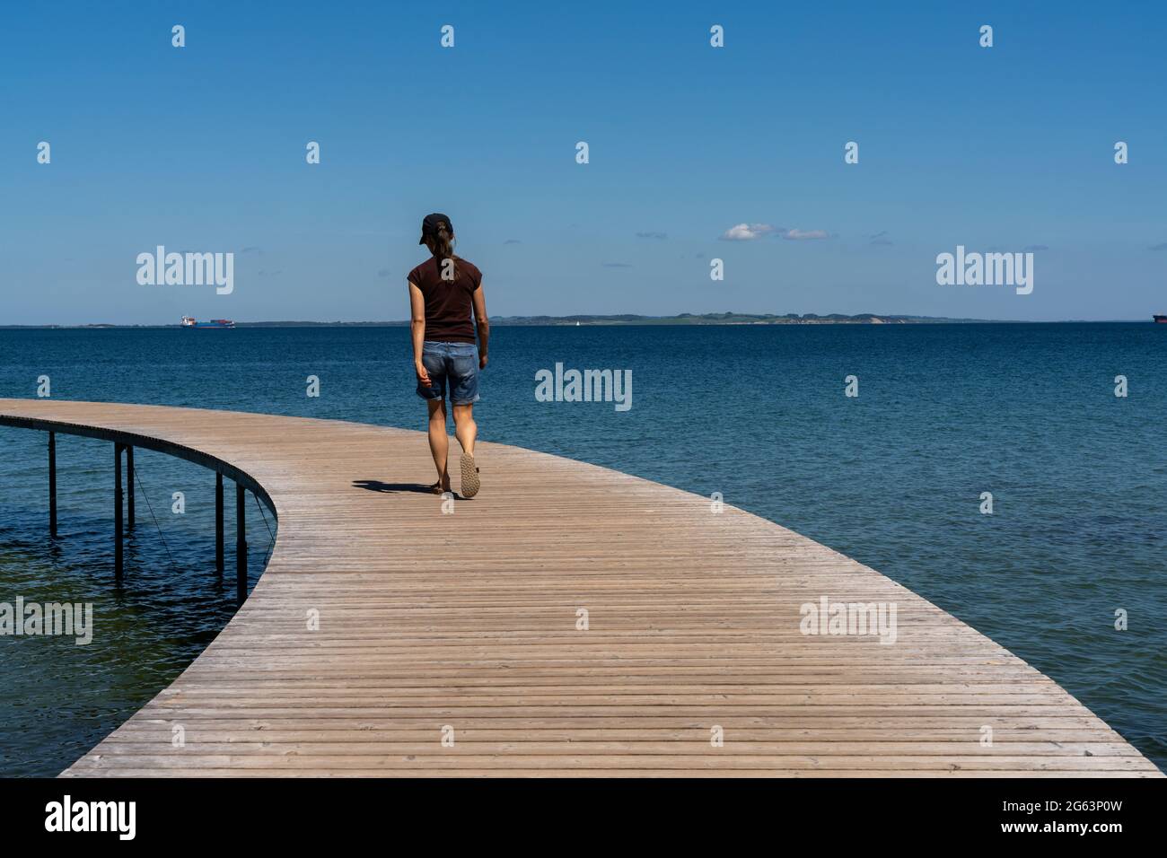 Eine schlanke Frau geht auf einer kreisförmigen Promenade entlang, die aus dem ruhigen, blauen Meer führt Stockfoto