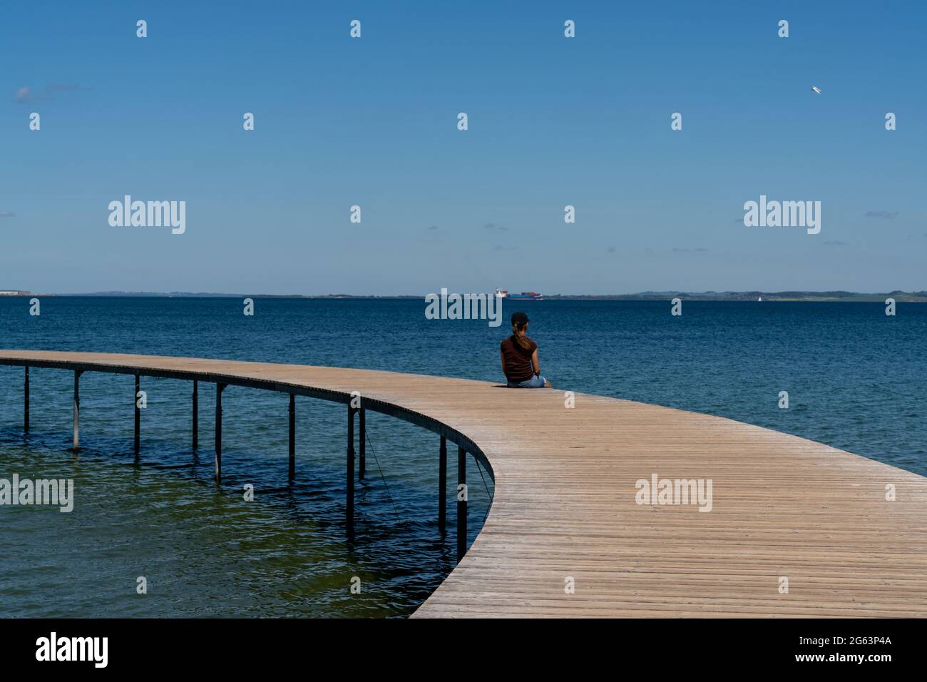 Eine schlanke Frau sitzt allein auf einer kreisförmigen Promenade, die über ein ruhiges, blaues Meer führt Stockfoto