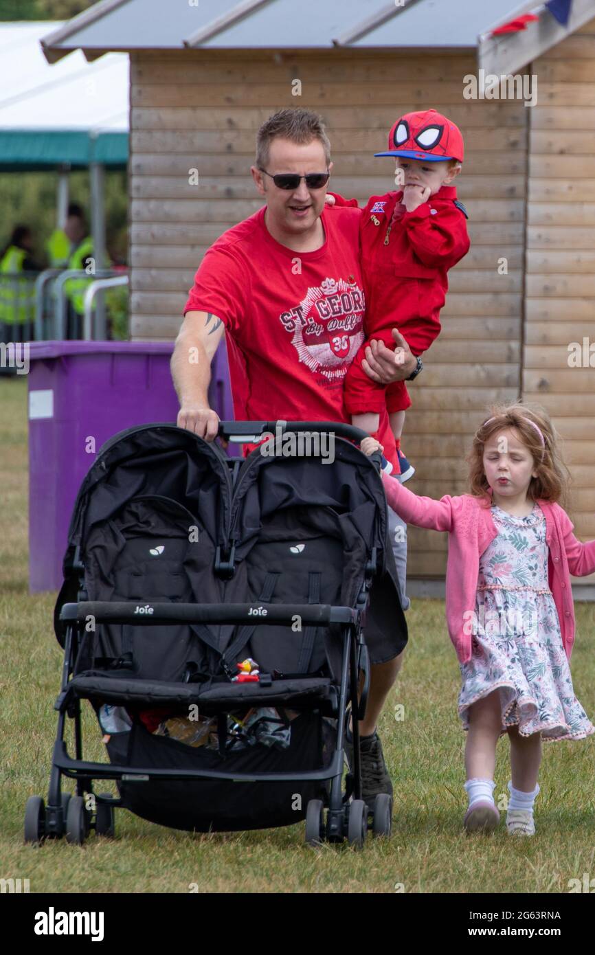 Ein Vater oder Vater schiebt einen Buggy Doppel-Buggy, während er ein Kind trägt, während das andere sich am Kinderwagen festhält Stockfoto