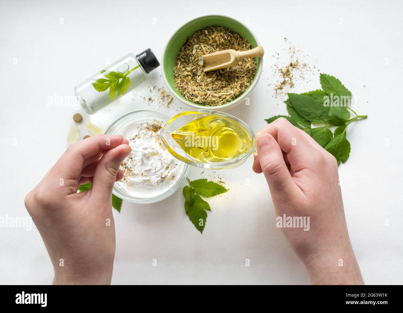 Vorbereitung einer alternativen Medizin, Kräuterextrakt: Weißer Hintergrund, Hände halten ein Glas Öl über eine Schüssel mit weißer Essenz, und Rühren. Schüssel mit g Stockfoto