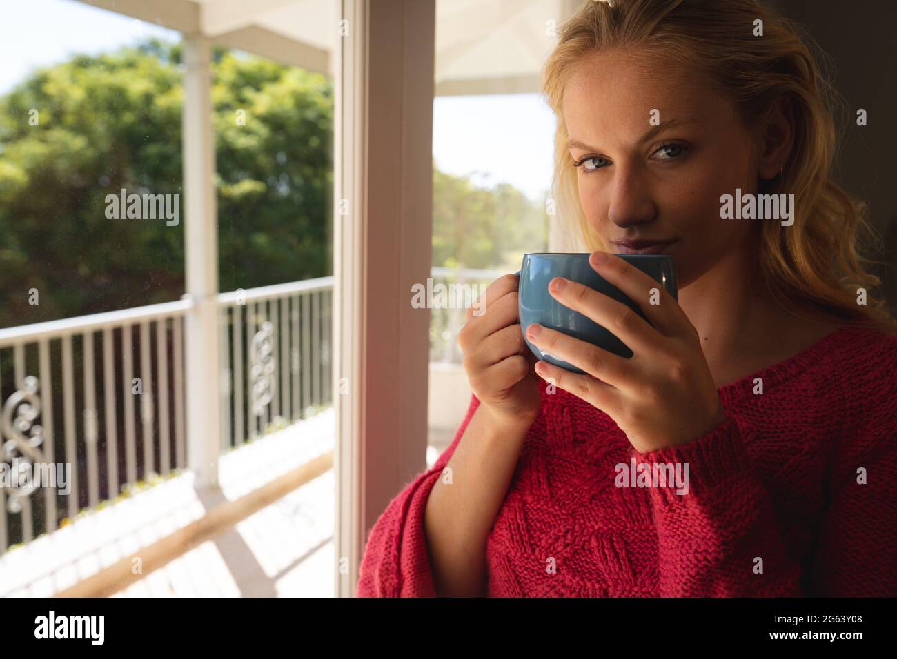 Porträt einer glücklichen kaukasischen Frau, die am sonnigen Balkonfenster steht und eine Tasse Kaffee hält Stockfoto