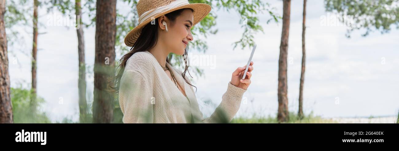 Positive junge Frau in Strohhut und schnurlosen Ohrhörern mit Smartphone im Wald, Banner Stockfoto