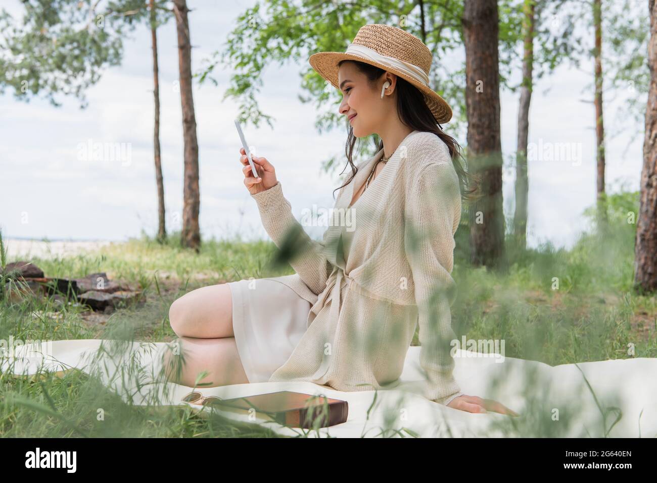 Lächelnde Frau mit Strohhut und schnurlosen Kopfhörern, die mit dem Smartphone auf einer Picknickdecke sitzt Stockfoto