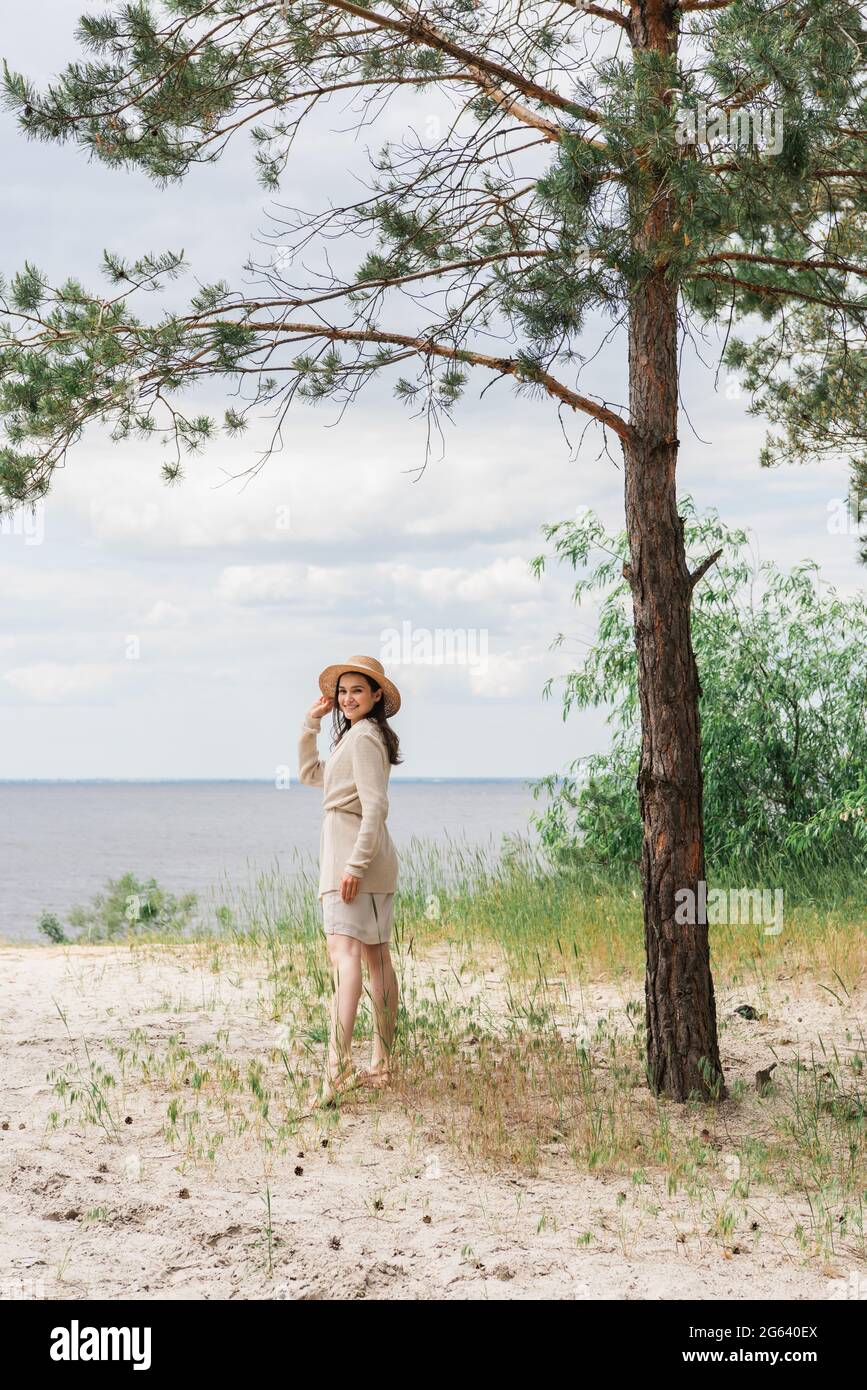 In voller Länge lächelnde junge Frau, die Strohhut anpasst und in Wäldern in der Nähe des Sees steht Stockfoto