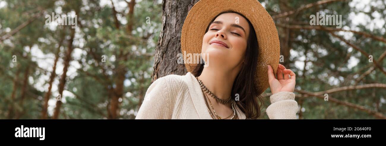 Fröhliche junge Frau, die Strohhut anpasst und im Wald lächelt, Banner Stockfoto